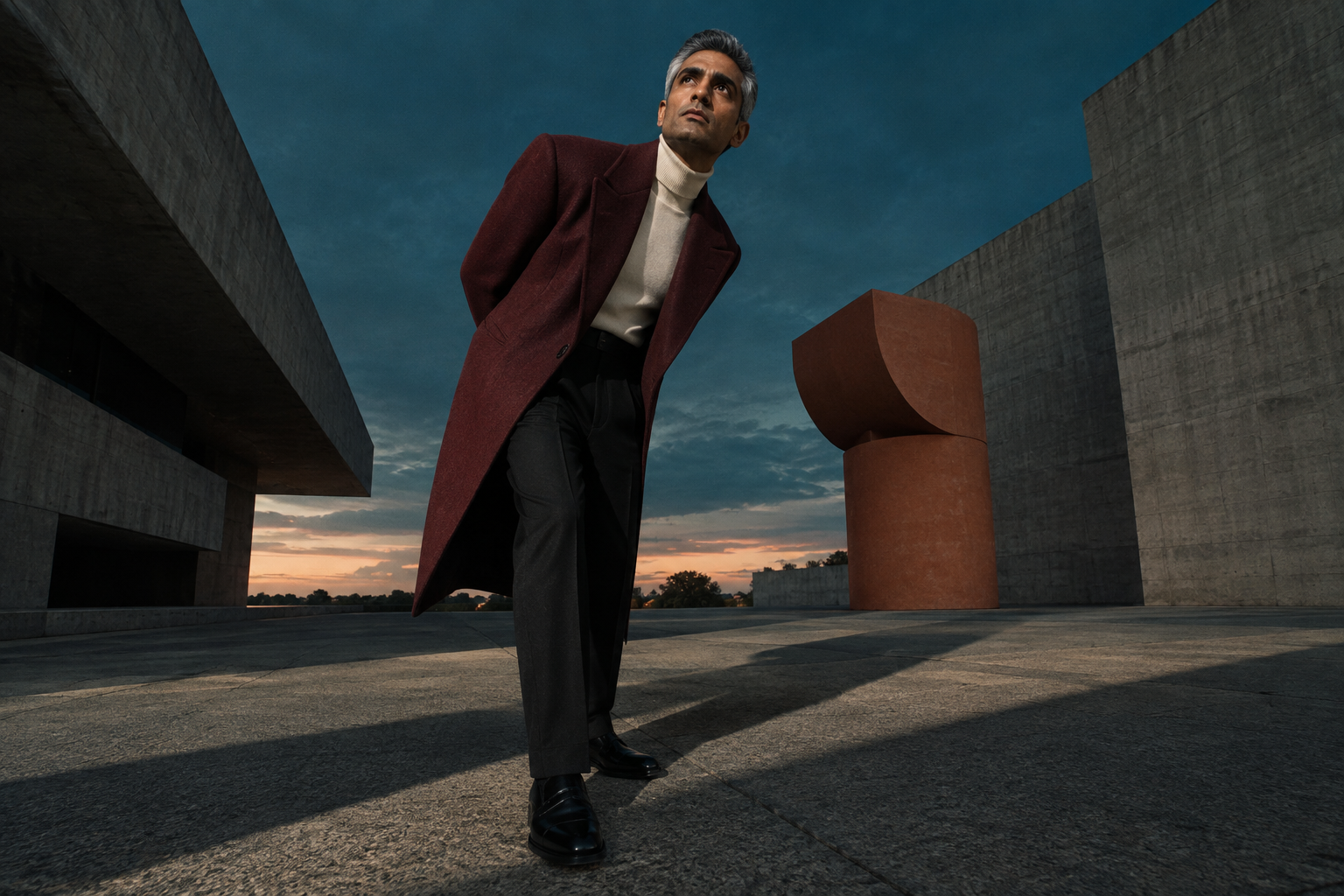 A stylish man posing in a long maroon coat against a backdrop of modern concrete buildings at dusk.