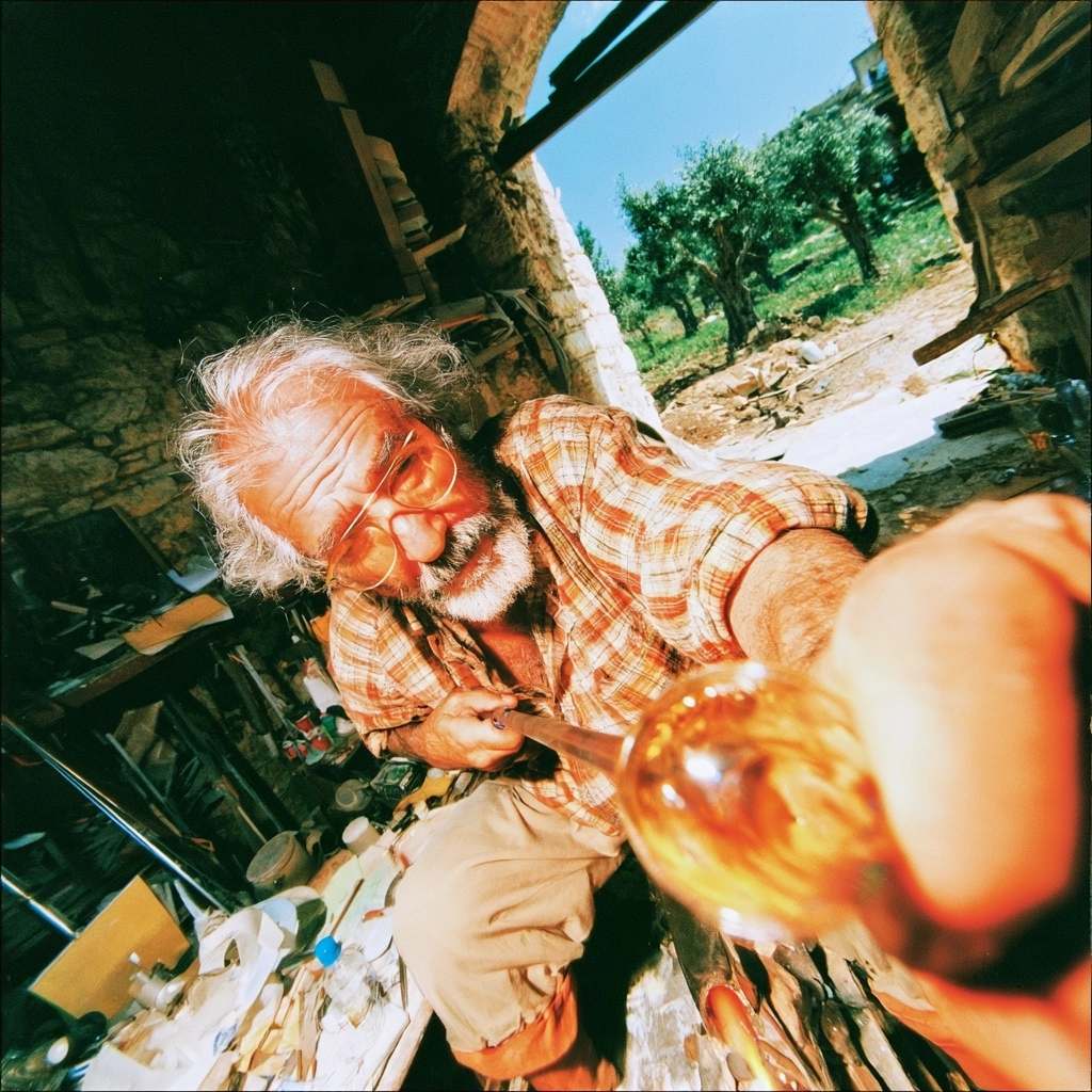 A bearded glassblower holding a glowing piece of molten glass in a stone workshop.