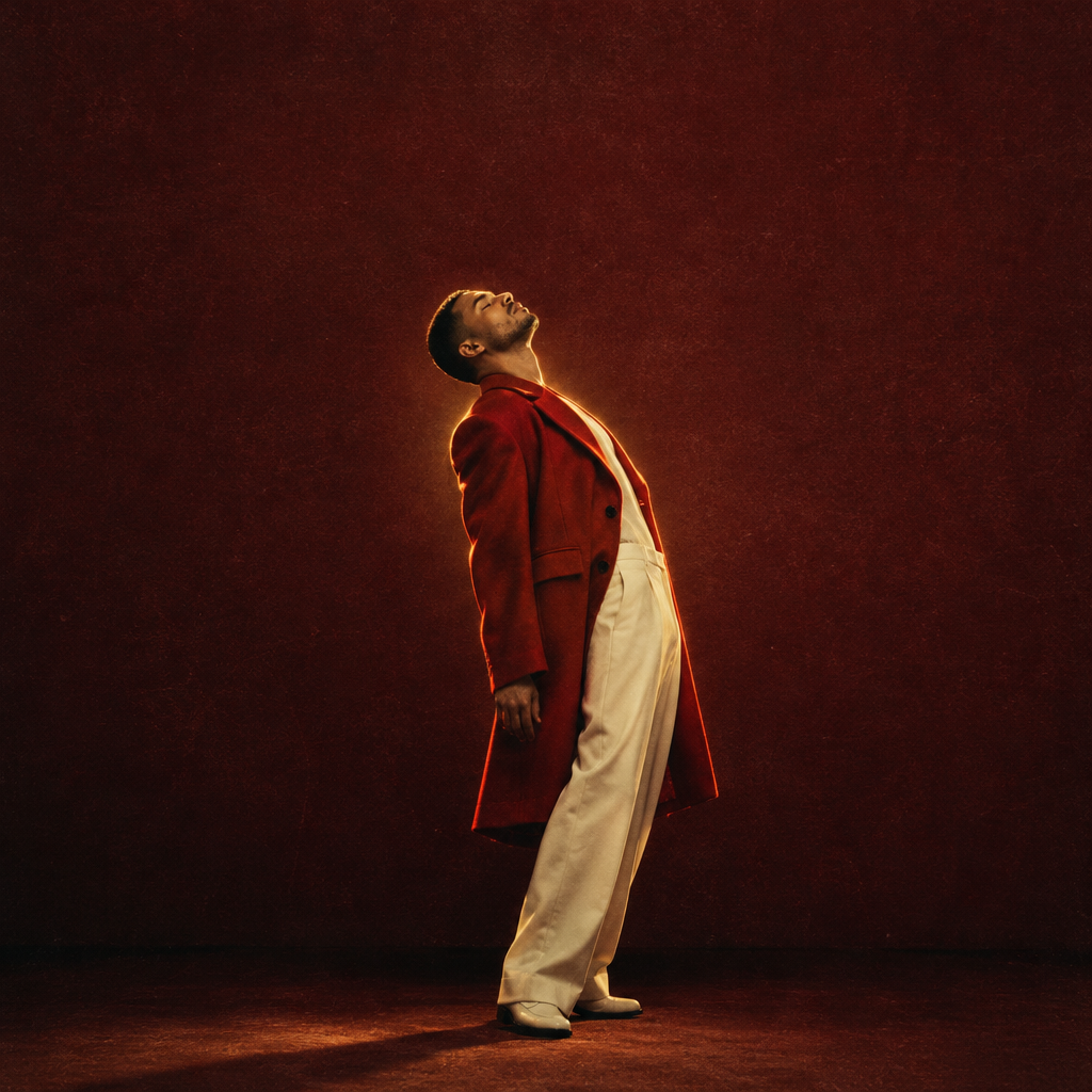 A man wearing a long red coat and white pants posing with his head tilted back against a red wall.
